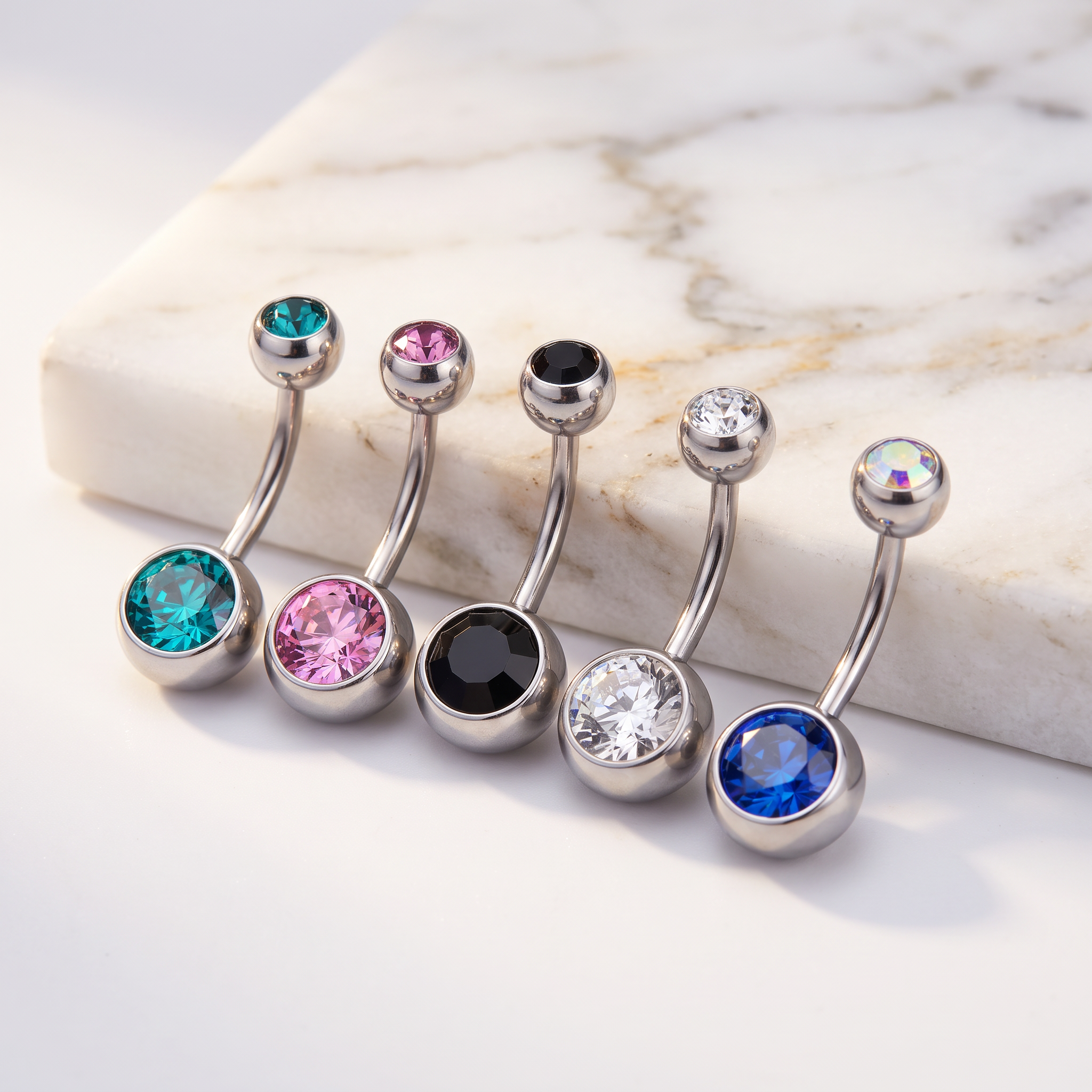 Five silver-toned belly button rings with round faceted gemstones in turquoise, pink, black, clear, and blue, arranged on a white marble slab against a white background.
