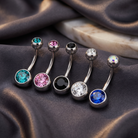 Five silver-toned curved barbell belly button rings with round faceted gemstones in turquoise, pink, black, clear, white, and blue, arranged on draped dark satin fabric.