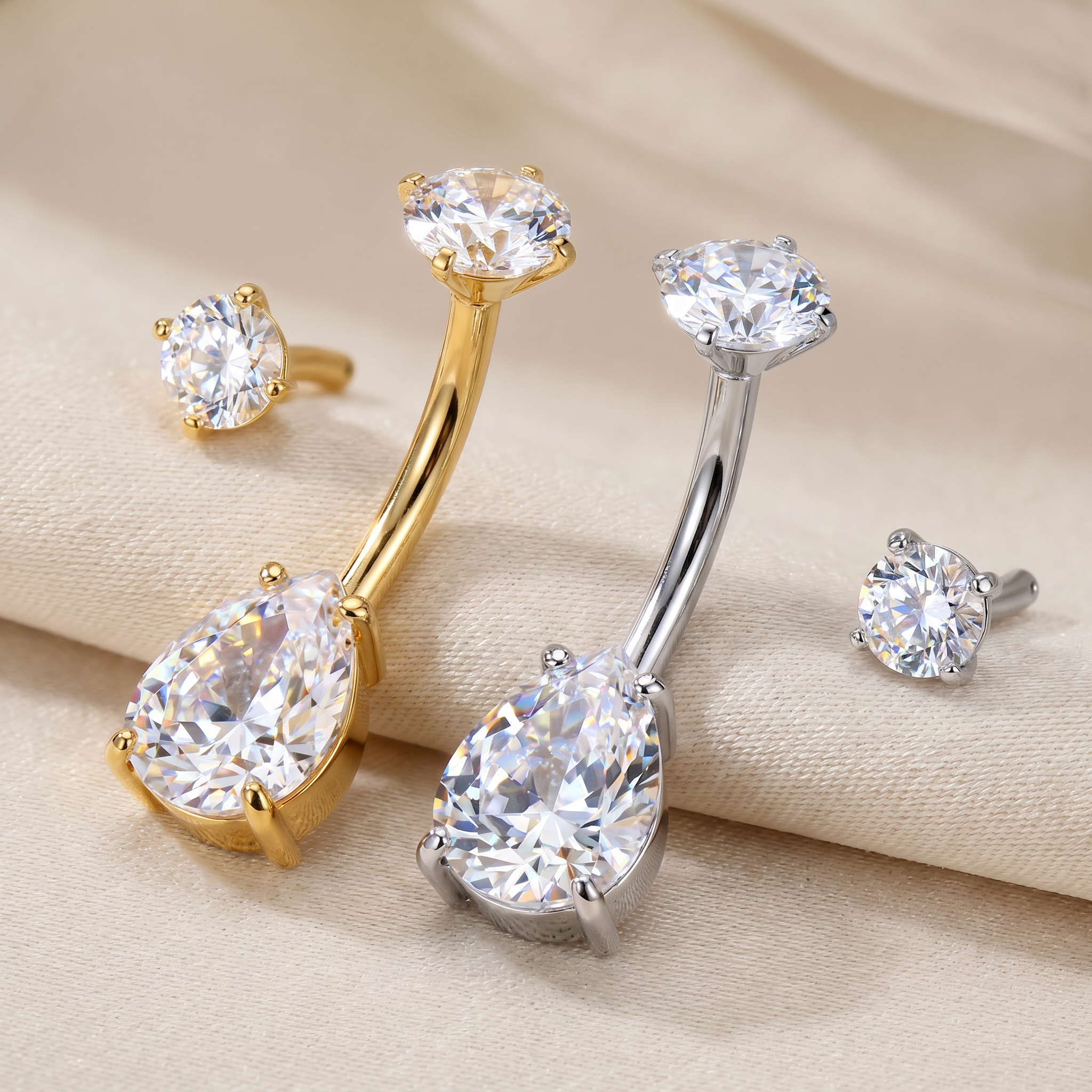 Two curved barbell belly button piercings with large teardrop-shaped clear gemstones, shown alongside matching round stud earrings on a beige fabric background.