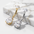 Two curved barbell belly button piercings with large teardrop-shaped clear gemstones, one in yellow gold and one in silver, displayed on a white marble block.