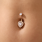 A close-up of a gold navel piercing with a small round stud above and a heart-shaped dangling charm below, both adorned with clear stones, set against human skin.