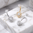 Four heart-shaped belly button piercings displayed on a white marble block, including two silver-toned pieces, one gold-toned piece, and one small matching stud, all featuring clear crystal rhinestone detailing.