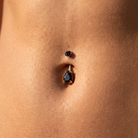 A close-up of a person's navel wearing a two-piece belly button jewelry set. The set includes a smaller top stud and a larger main ring with a teardrop-shaped black gemstone in a gold setting, connected by a straight gold bar.