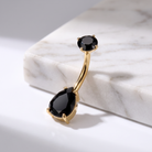 Gold curved barbell navel piercing jewelry with two black faceted gemstones, displayed on a white marble block.