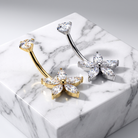Two flower-shaped belly button rings with clear marquise-cut gemstones, one in yellow gold and one in silver, displayed on a white marble block against a light gray background.