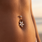 A close-up of a person's lower torso wearing a gold belly button ring with a flower-shaped crystal setting and a smaller matching stud above it.