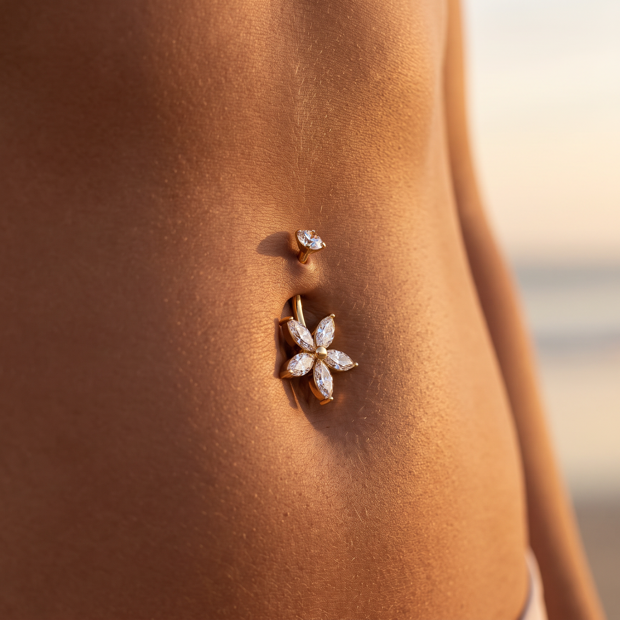 A close-up of a person's lower torso wearing a gold belly button ring with a flower-shaped crystal setting and a smaller matching stud above it.