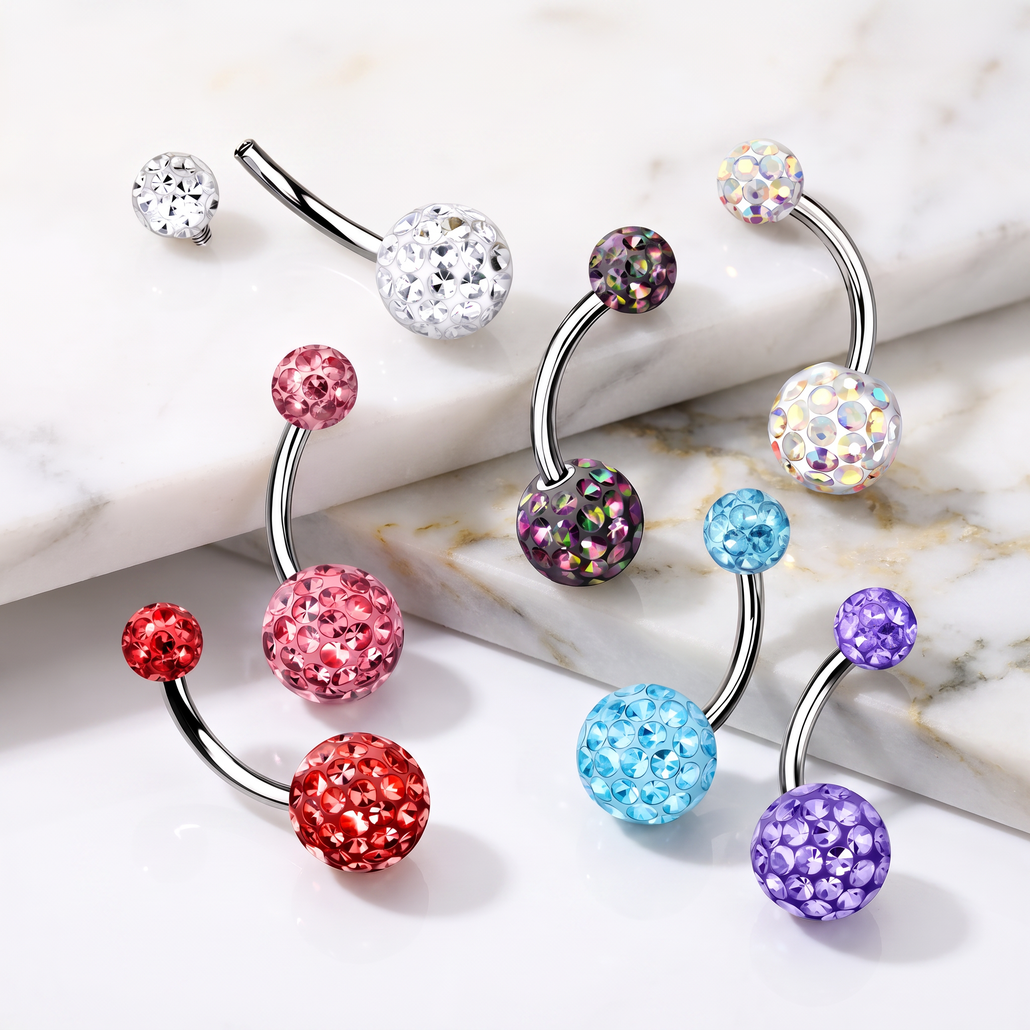 Four curved barbell belly button piercings with sparkling rhinestone-covered balls in assorted colors, arranged on a white marble surface.