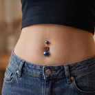 Close-up of a person's lower torso wearing blue jeans and a black crop top, showing a blue metallic navel piercing with two small blue studs.
