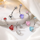 Five silver-toned belly button rings with heart-shaped crystal studs in red, clear, purple, pink, and light blue, arranged on a white marble slab with white satin fabric.