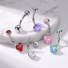 Six silver-toned belly button ring barbells arranged on a white marble slab and white surface, each decorated with a heart-shaped crystal in a different color: red, clear, purple, pink, blue, and clear.