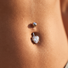 A close-up of a person's lower abdomen showing a silver-toned belly button ring with a heart-shaped clear gemstone and a smaller round silver bead above it.