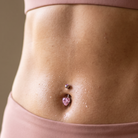Close-up of a person's midsection wearing a dusty pink sports bra and matching leggings, with a pink gem belly button ring visible at the navel.