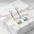 Two curved barbell belly button piercings with rectangular clear gemstones, shown on a white marble display block. One piece is gold-toned and the other is silver-toned.