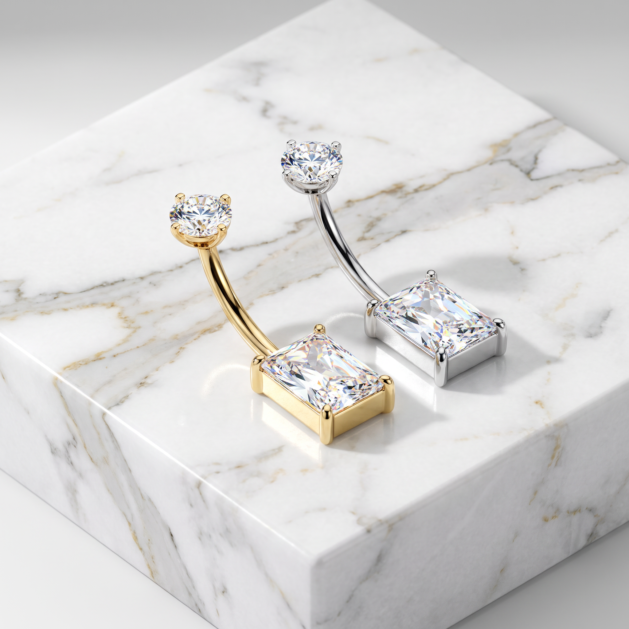 Two curved barbell belly button piercings with rectangular clear gemstones, shown on a white marble display block. One piece is gold-toned and the other is silver-toned.