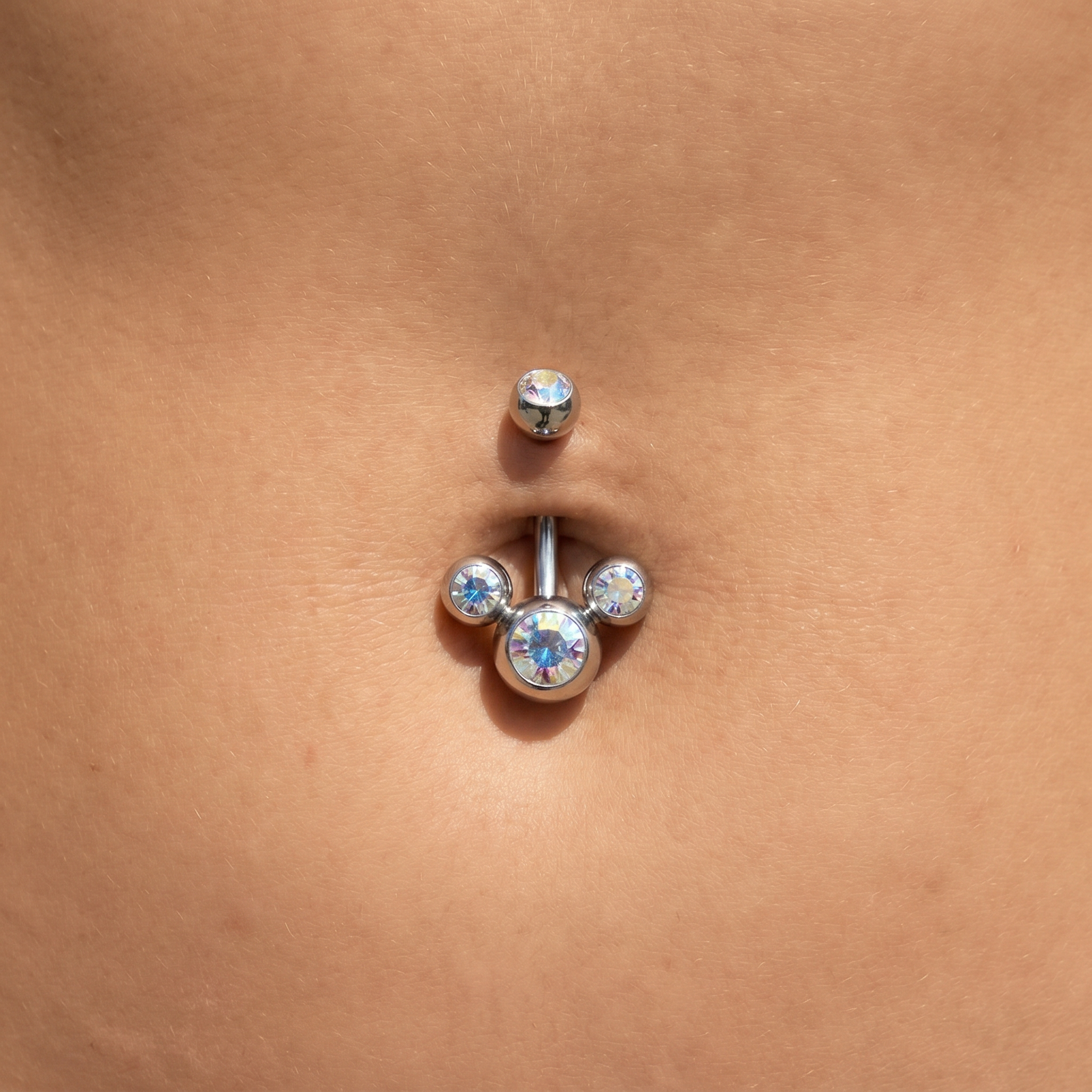 A close-up of a silver-colored navel piercing with a curved barbell and clear rhinestone accents, shown on a person's abdomen.
