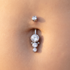 A close-up of a silver belly button ring with three clear round gemstones, worn on a person's navel.