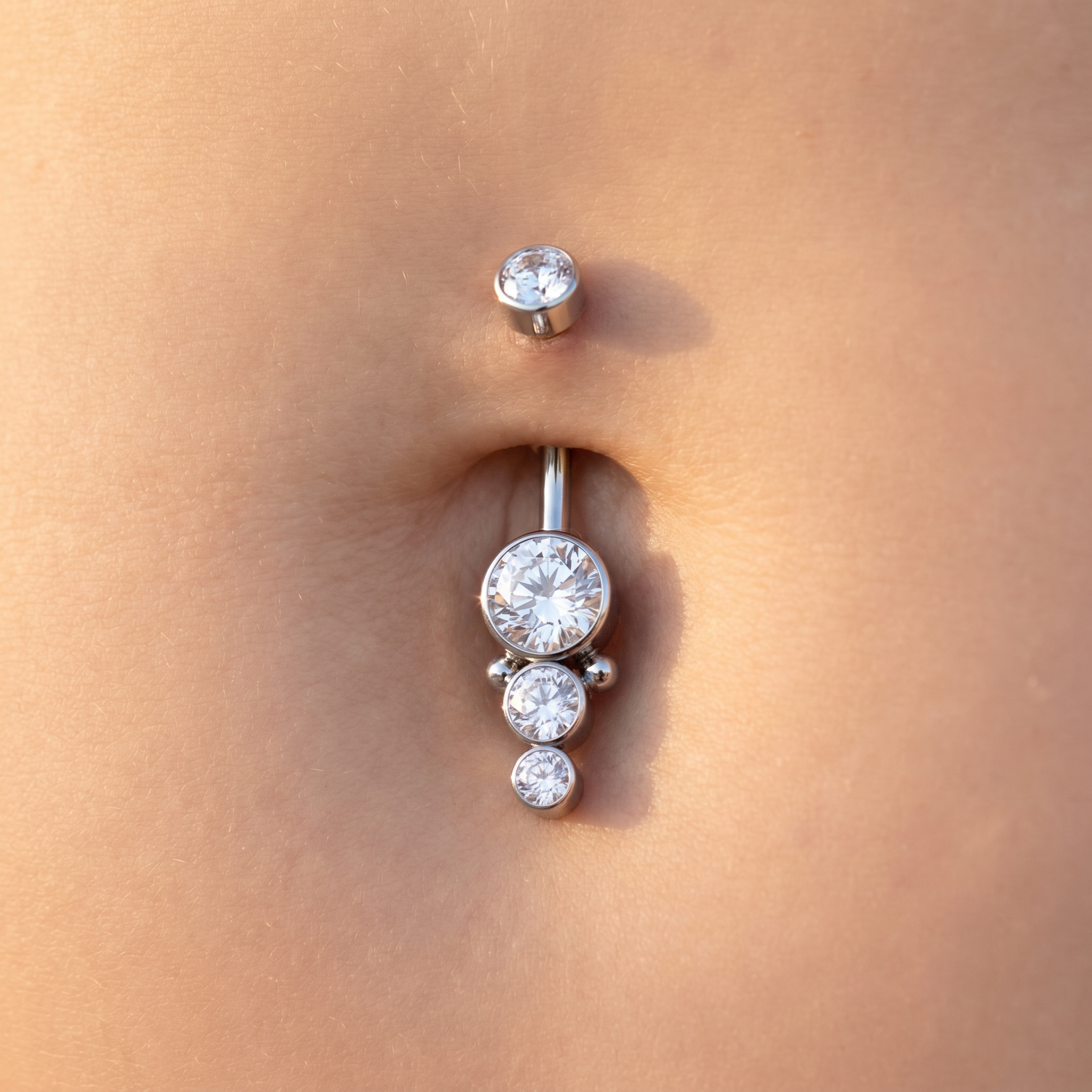A close-up of a silver belly button ring with three clear round gemstones, worn on a person's navel.