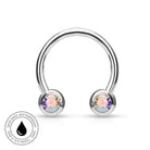 A silver-toned curved barbell piercing stud with two round iridescent gemstones, shown on a white background with a circular 'Skin Safe Waterproof and Tarnish Free' badge in the lower left.