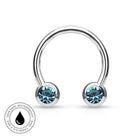 A silver-colored curved barbell piercing stud with two round bezel-set light blue gemstones, shown on a white background with a circular 'Skin Safe Waterproof' badge in the bottom-left corner.