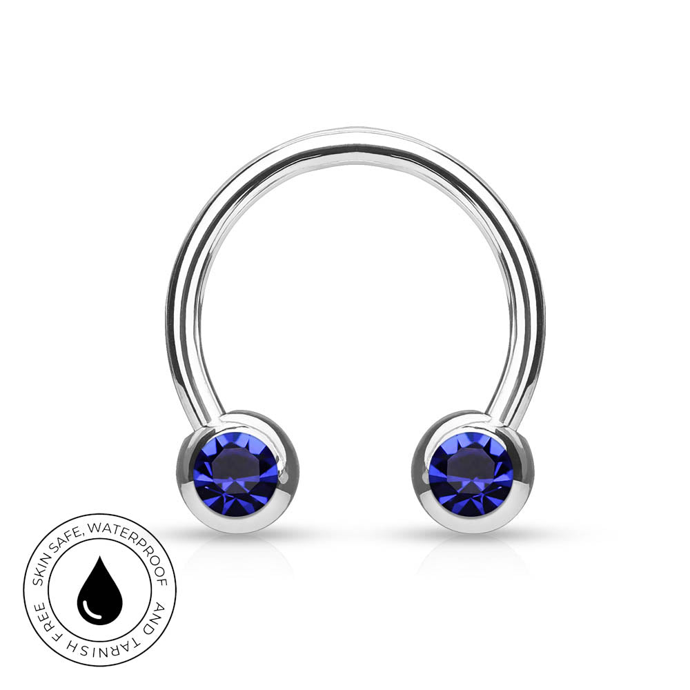 A silver-toned curved barbell piercing stud with two round deep blue gemstones, shown on a white background with a circular 'Skin Safe Waterproof' badge in the lower-left corner.