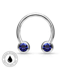 A silver-toned curved barbell piercing stud with two round deep blue gemstones, shown on a white background with a circular 'Skin Safe Waterproof' badge in the lower-left corner.