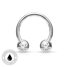 A silver-toned curved barbell piercing stud with two clear faceted gemstones in round bezel settings, shown on a white background with a circular 'skin-safe waterproof' badge in the lower-left corner.