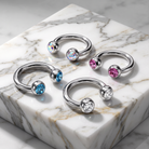 Four silver-toned hoop-style body jewelry pieces with round faceted gemstones in iridescent, blue, pink, and clear settings, arranged on a white marble block.