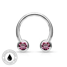 A silver-toned curved barbell piercing stud with two round pink gemstones, shown on a white background with a circular 'Skin Safe Waterproof' badge in the lower left corner.