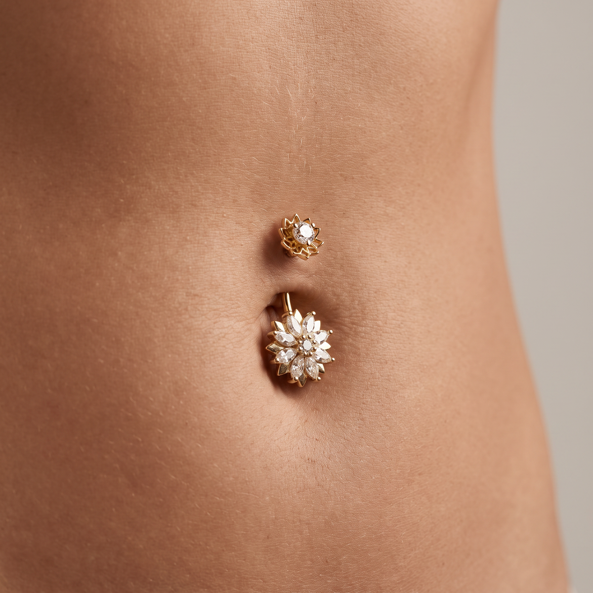 A close-up of a person's lower torso showing two gold belly button piercings: a small floral stud above and a larger matching flower navel ring below.