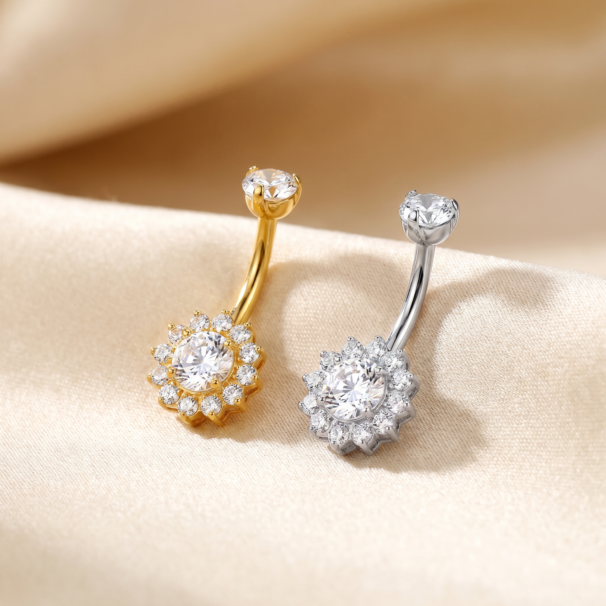 Two flower-shaped belly button rings with clear gemstones, one in yellow gold and one in silver, displayed on a beige satin fabric background.