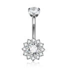 A silver-toned navel piercing ring with a single clear gemstone at the top and a flower-shaped cluster of small clear gemstones at the bottom. An inset circle in the upper right shows the threaded end of the barbell.