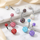 Four curved barbell belly button piercings with sparkling rhinestone-covered balls in assorted colors, arranged on a light marble surface with soft cream fabric in the background.