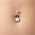 A close-up of a belly button with a silver-toned navel piercing. The lower stud features a large, round, iridescent white cabochon gemstone, while the upper stud has a smaller, similar round gemstone.