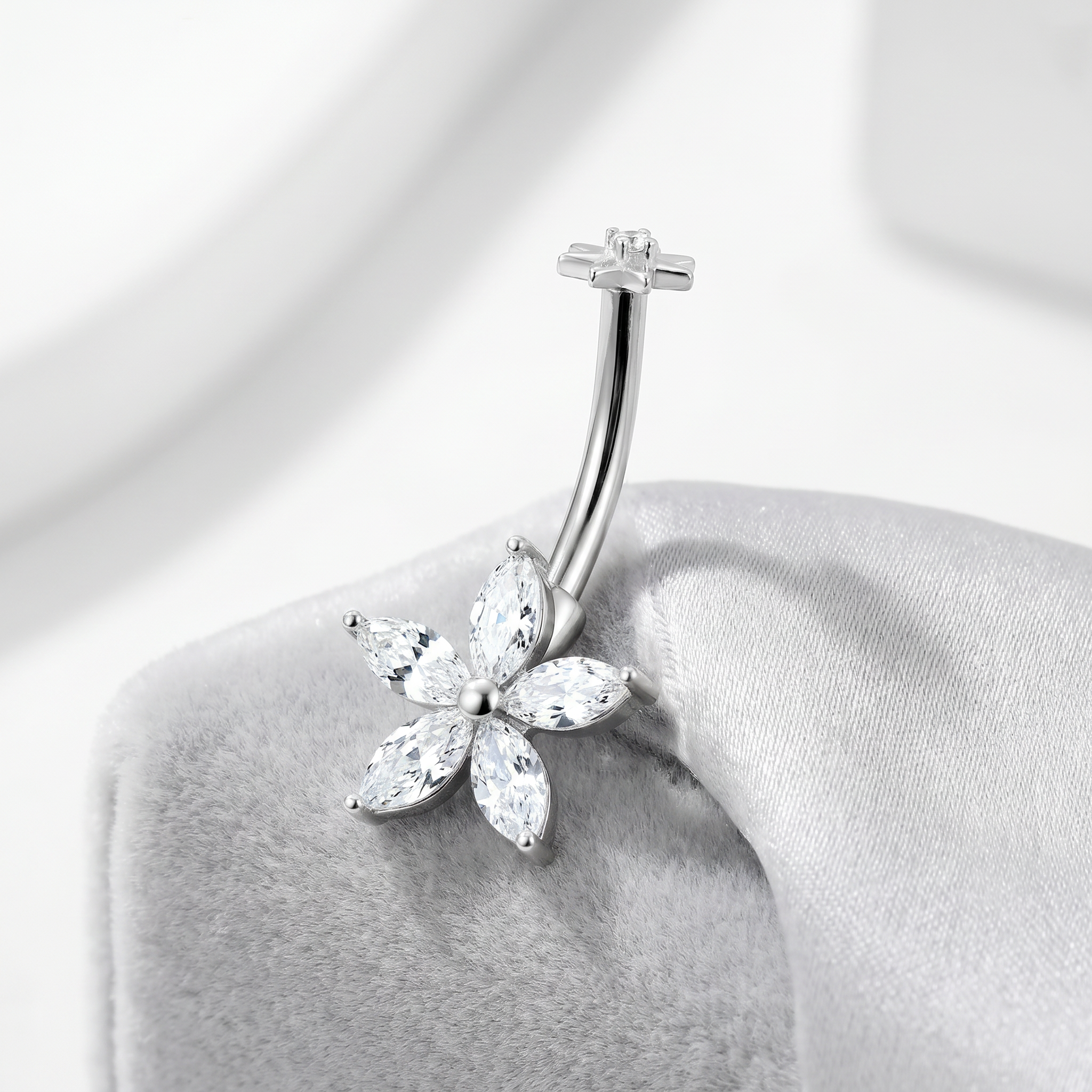 A silver-toned curved barbell belly button ring with a five-petal flower setting made of clear marquise-cut stones, displayed on a soft gray jewelry stand.