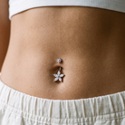 A close-up of a person's lower torso wearing a silver belly button ring with a flower-shaped charm and a small matching stud, paired with a white ribbed crop top and white drawstring shorts.