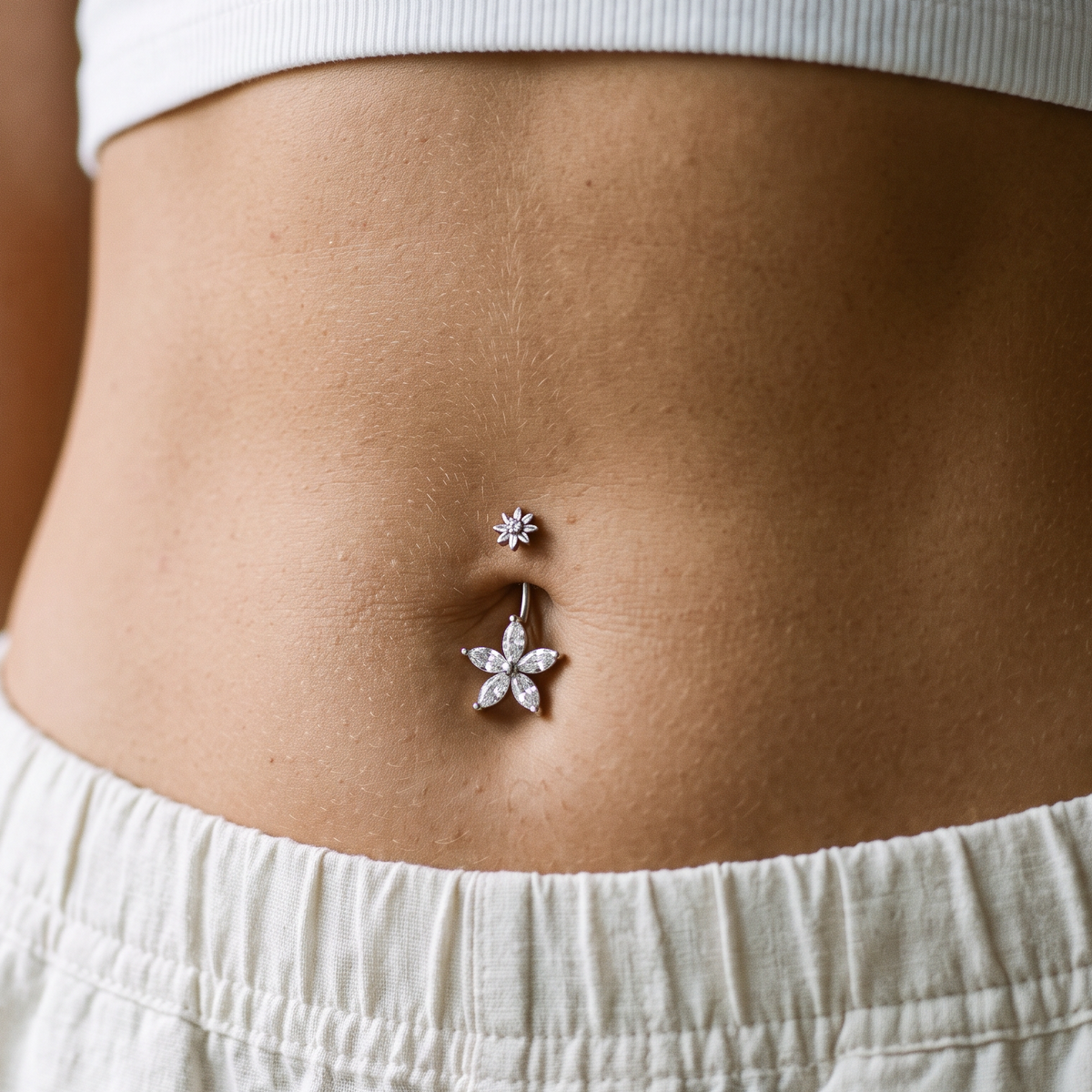 A close-up of a person's lower torso wearing a silver belly button ring with a flower-shaped charm and a small matching stud, paired with a white ribbed crop top and white drawstring shorts.