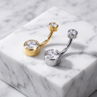 Two curved barbell belly button piercings displayed on a white marble block. One is gold-toned with a large clear gemstone, and the other is silver-toned with a matching large clear gemstone.