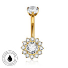 Gold-plated navel ring with a single round clear gemstone on the top and a flower-shaped halo of smaller clear gemstones around a central round clear gemstone on the bottom, shown on a white background with a small circular 'skin-safe waterproof' badge in the lower left.