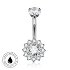 Silver-toned navel ring with a single round clear gemstone at the top and a flower-shaped halo of smaller clear gemstones at the bottom, shown on a white background with a circular skin-safe waterproof badge in the lower left.