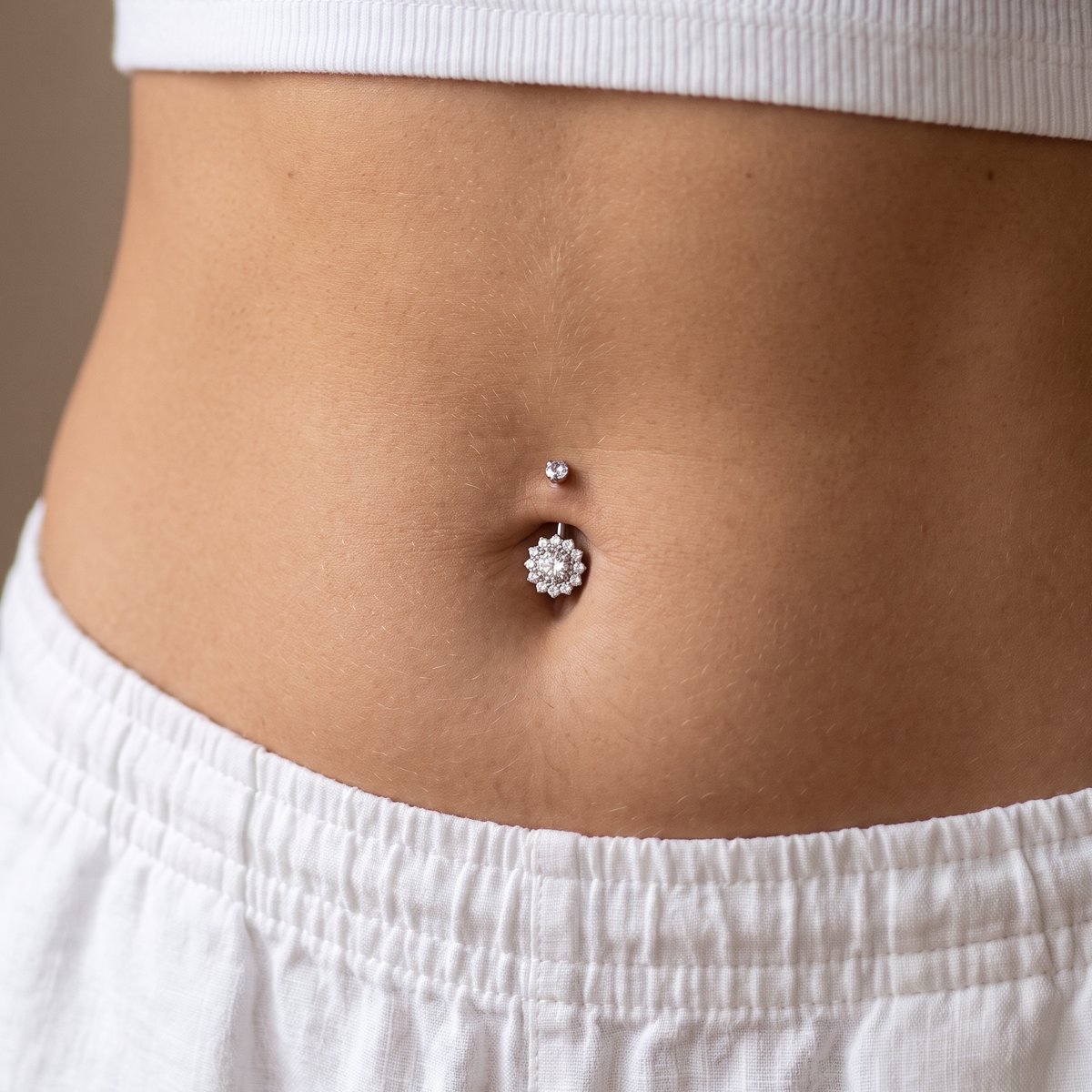 A close-up of a person's lower torso showing a belly button piercing with a small silver stud and a larger silver flower-shaped stud, worn with a white ribbed crop top and white shorts.