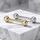 Two barbell stud earrings displayed on a white marble block. One earring is gold-toned with a clear gemstone, and the other is silver-toned with a clear gemstone.
