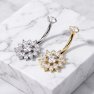 Two flower-shaped belly button rings with clear cubic zirconia stones, one in silver and one in gold, displayed on a white marble block.