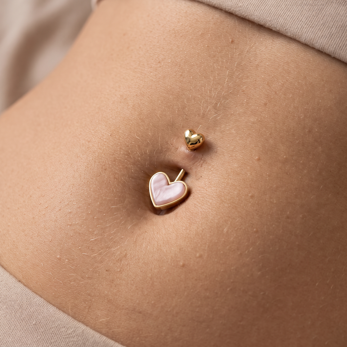 A close-up of a person's navel area showing two belly button piercings: a larger gold heart stud with a pale pink inlay and a smaller gold heart stud above it, against light beige fabric.