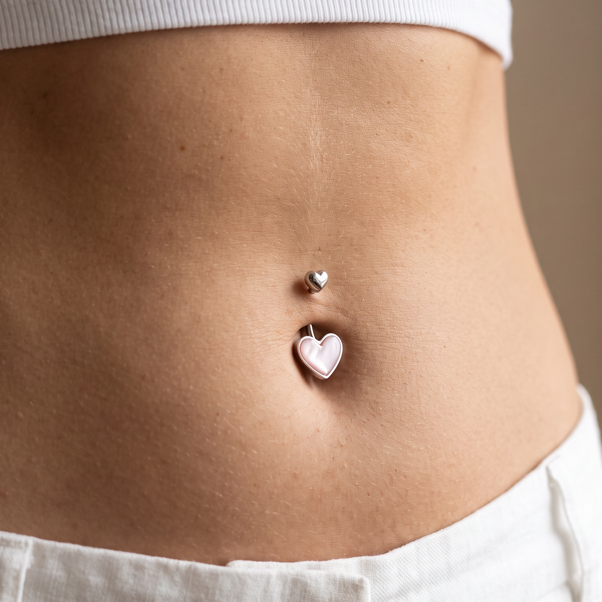 A close-up of a person's midsection wearing a silver belly button ring with a small heart stud and a larger dangling heart charm.