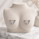 A pair of silver-toned crescent nipple piercings with white pearl centers and clear rhinestone accents, displayed on a beige jewelry bust.