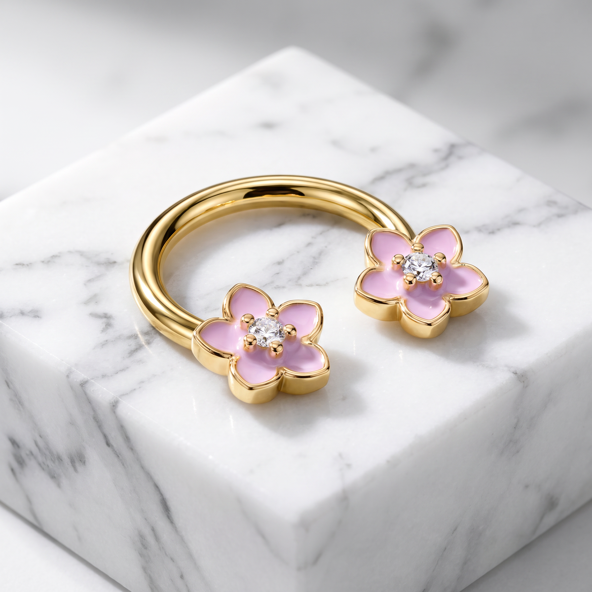 A gold-plated open hoop ring decorated with two pink enamel flower charms, each featuring a clear round gemstone in the center, displayed on a white marble block.