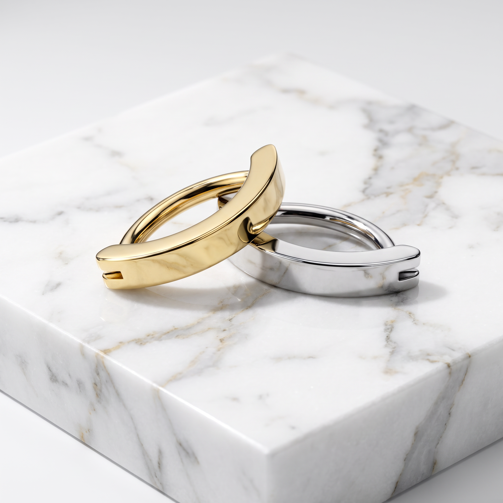 Two minimalist curved hoop-style rings, one in polished yellow gold and one in polished silver, displayed on a white marble block against a light gray background.