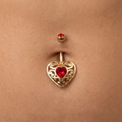 Gold belly button ring with an ornate filigree heart pendant featuring a red heart-shaped gemstone, shown worn on a person's navel.