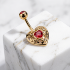 Gold-plated belly button ring with an ornate filigree heart design and a red heart-shaped gemstone, resting on a white marble cube.